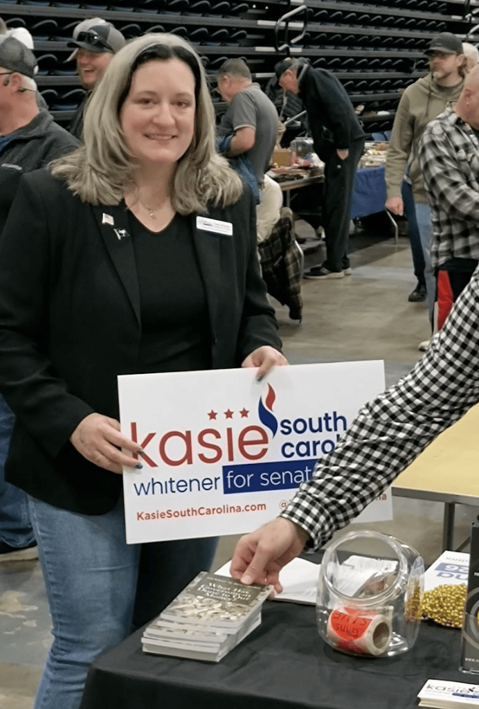 Kasie stands with a campaign sign at a table at the gun show